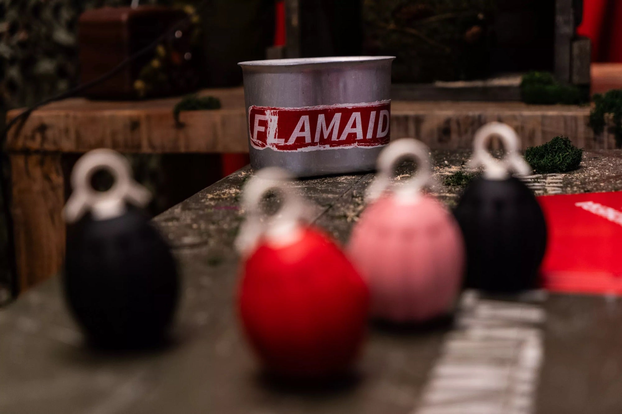 Granadas FlamAid en negro, rojo y rosa sobre una mesa, con una lata serigrafiada FlamAid al fondo